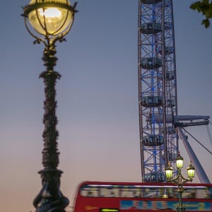 May include: A nighttime cityscape featuring a decorative black lamppost with a glowing light, a red double-decker bus, and the London Eye Ferris wheel against a dusky blue sky. The Ferris wheel is illuminated with blue lights.