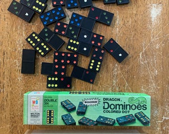 Puede incluir: Un conjunto de dominós negros con puntos de colores dispersos sobre una superficie de madera. También hay una caja verde y blanca de "Dragon Dominoes" de Milton Bradley, junto con una caja de cartón.
