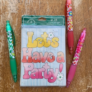 May include: A set of party-themed stationery on a wooden surface. The set includes a package of cards with the text "Let's Have a Party!" and four pens with colorful designs. The pens have red, pink, and green accents.