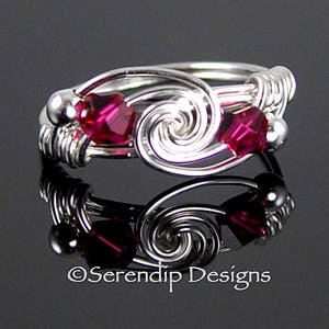 May include: A silver wire wrapped ring with two pink crystal beads. The ring features a swirling design.