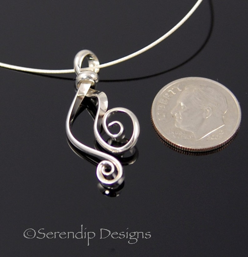 May include: A silver-colored pendant in the shape of a treble clef, with a spiral design. The pendant is on a thin, silver-colored necklace. A U.S. dime is next to the pendant for size comparison. The pendant is on a black surface.
