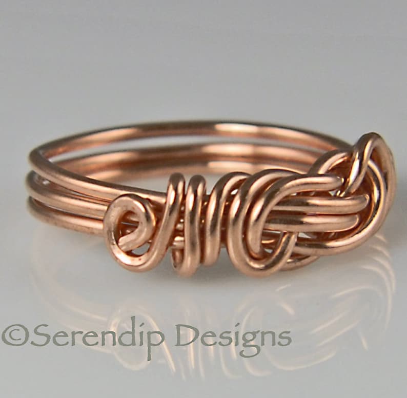 Puede incluir: Un anillo artesanal de alambre de cobre con un dise&ntilde;o &uacute;nico tejido. El anillo presenta m&uacute;ltiples bucles y espirales, creando una apariencia intrincada y art&iacute;stica. El anillo es de color cobre c&aacute;lido y se muestra sobre un fondo claro.