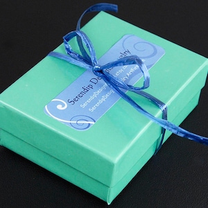 Puede incluir: Una peque&ntilde;a caja de regalo turquesa atada con una cinta azul. Una etiqueta azul y blanca en la caja dice "Serendip Designs". La caja est&aacute; cerrada y lista para regalar. El fondo es negro.
