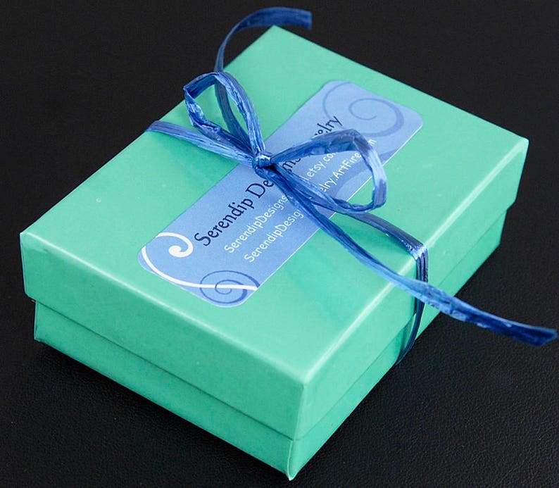 Puede incluir: Una peque&ntilde;a caja de regalo turquesa atada con una cinta azul. Una etiqueta azul y blanca en la caja dice "Serendip Designs". La caja est&aacute; cerrada y lista para regalar. El fondo es negro.