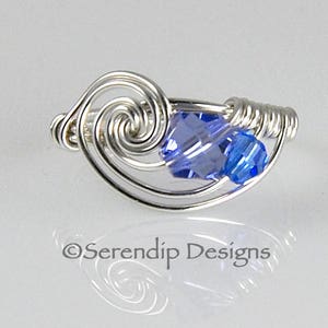 Può includere: Un anello in filo d'argento con un cristallo blu. Il filo è attorcigliato a forma di spirale. L'anello è un gioiello delicato.
