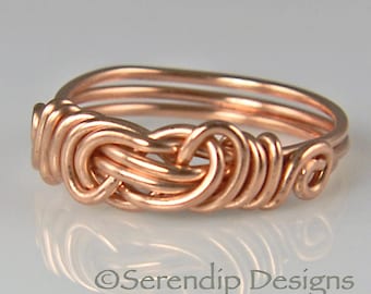 May include: A copper wire ring with a woven knot design. The ring is made from copper wire, with a series of looped wires creating a textured appearance. The ring is a warm copper colour and has a polished finish. The text "Serendip Designs" is at the bottom.