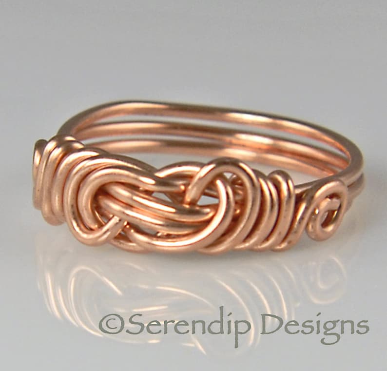 Puede incluir: Un anillo artesanal de alambre de cobre con un dise&ntilde;o de nudo tejido. El anillo presenta una serie de alambres en bucle, creando una apariencia intrincada y texturizada. El anillo es de color cobre c&aacute;lido y tiene un acabado pulido. El texto "Serendip Designs" est&aacute; en la parte inferior.