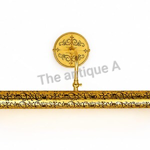 May include: A gold-toned metal picture light featuring a cylindrical shade with an intricate black filigree pattern. The fixture includes a circular wall-mounting plate with matching black scrollwork and the text The antique A.