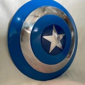 May include: A circular shield featuring a vibrant blue outer ring, a polished silver metallic middle ring, and a dark blue center circle. A five-pointed silver star is centered within the dark blue circle, displaying visible rivets at each point.