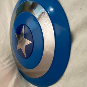 May include: A circular shield featuring a glossy blue outer ring, a reflective silver middle ring, and a central blue circle containing a five-pointed silver star. The design is concentric, with smooth, curved surfaces and a metallic finish.