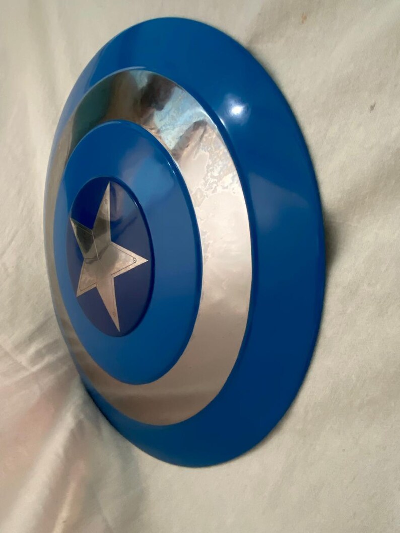 May include: A circular shield featuring a glossy blue outer ring, a reflective silver middle ring, and a central blue circle containing a five-pointed silver star. The design is concentric, with smooth, curved surfaces and a metallic finish.
