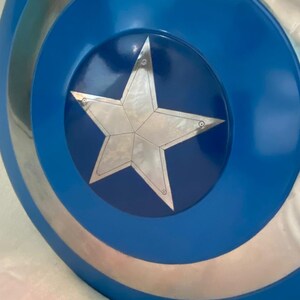 May include: A circular shield featuring a metallic silver star at the center, set against a dark blue circular background. The outer edge of the shield consists of alternating concentric rings of metallic silver and vibrant blue, creating a distinct pattern.