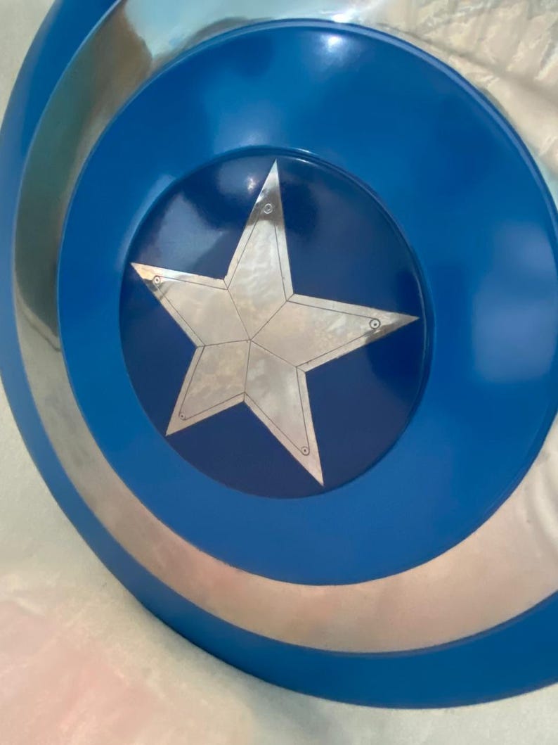 May include: A circular shield featuring a metallic silver star at the center, set against a dark blue circular background. The outer edge of the shield consists of alternating concentric rings of metallic silver and vibrant blue, creating a distinct pattern.