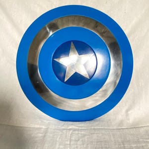 May include: A circular shield featuring a bright blue outer ring, a metallic silver middle ring, and a central blue circle containing a prominent five-pointed silver star. The design is symmetrical with a smooth, reflective finish throughout the surface.