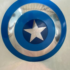 May include: A circular shield featuring a vibrant blue outer ring, a reflective silver middle ring, and a dark blue center circle. A prominent five-pointed silver star is centered within the dark blue circle, creating a bold, geometric design.