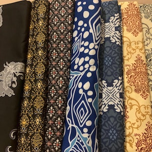 以下が含まれることがあります： さまざまな模様と色を持つ、丸められた生地のコレクション。デザインには、黒、金、青、クリーム色の花柄、幾何学模様、抽象的なモチーフが含まれます。生地は綿または同様の素材で、縫製や手芸プロジェクトに適しているようです。