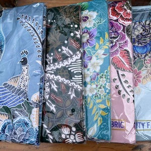 Puede incluir: Rollos de tela con coloridos estampados florales y de pavo real. Las telas presentan tonos de azul, verde, rosa y morado. Algunos rollos tienen las palabras "BRIC" y "TY FABRIC" impresas.