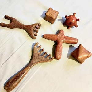 Può includere: Una collezione di strumenti per massaggio in legno fatti a mano in varie forme. Include uno strumento a forma di rastrello, un massaggiatore a forma di croce, una palla spinosa e forme geometriche. Gli strumenti sono in legno marrone rossiccio.