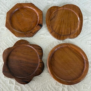 Könnte beinhalten: Vier Untersetzer aus Holz in verschiedenen Formen: ein Achteck, ein abgerundetes Quadrat, eine Blume und ein Kreis. Die Untersetzer bestehen aus warmbraunem Holz mit sichtbaren Maserungsmustern. Sie schützen Oberflächen vor Kondensation.