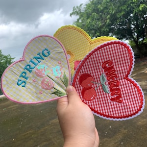 以下が含まれることがあります： 3つのハート型の布パッチ。1つは黄色のギンガムチェックで、ターコイズブルーで「SPRING」と刺繍され、花のアクセントが付いています。もう1つは赤と白のギンガムチェックで「CHERRY」と刺繍されています。3つ目は、スカラップエッジの黄色です。