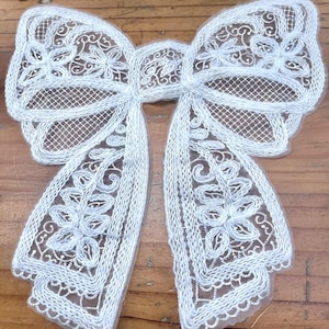 Puede incluir: Un lazo de encaje blanco con intrincados detalles florales y de volutas. El lazo presenta un diseño delicado y calado con largas colas fluidas. El encaje se coloca sobre un fondo de madera, resaltando la delicada textura y el diseño del lazo.