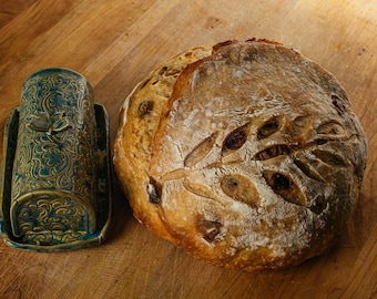 May include: A rustic loaf of bread with decorative cuts and a teal ceramic butter dish. The bread is golden brown. The butter dish has a floral design and a butterfly accent. The bread and butter dish are on a wooden surface.
