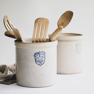 Może przedstawiać: Dwa cylindryczne, kremowobiałe ceramiczne pojemniki z niebieską pieczątką w kształcie żołędzia z napisem Uhl Pottery Co. Wares. Przedni pojemnik zawiera kilka jasnych drewnianych przyborów kuchennych, w tym łopatkę z otworami, łyżkę i szpatułkę.