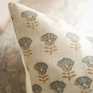 May include: A beige throw cushion cover featuring a repeating pattern of small, stylised flowers with light blue petals and brown stems. The fabric has a visible woven texture, and the cushion is shown at an angle with a white side seam.