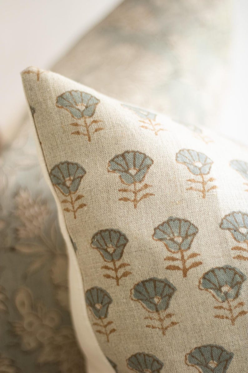May include: A beige throw cushion cover featuring a repeating pattern of small, stylised flowers with light blue petals and brown stems. The fabric has a visible woven texture, and the cushion is shown at an angle with a white side seam.