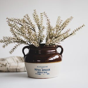 May include: A vintage brown and cream-colored stoneware crock with two handles, filled with white floral stems. The crock has text that reads "Compliments of Peter Schuck, General Store, Zillinger, Wis."
