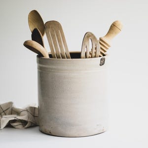 May include: A cylindrical light gray ceramic crock filled with various wooden kitchen utensils, including spoons, a slotted turner, and a ridged rolling pin. A beige cloth with a thin black grid pattern is positioned to the left of the crock.