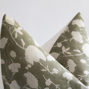 Puede incluir: Dos fundas de coj&iacute;n con estampado verde y blanco con un borde blanco. El estampado presenta un dise&ntilde;o floral con flores y hojas blancas sobre un fondo verde.