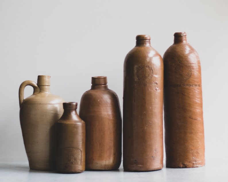 May include: A selection of antique stoneware bottles and jugs in shades of brown. The collection includes a jug with a handle, several bottles with rounded bodies, and a smaller bottle with a label. The vessels have a rustic, aged appearance, typical of vintage items.