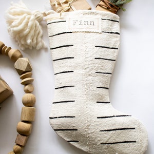May include: A cream-colored fabric Christmas stocking featuring horizontal black stripes and a white yarn tassel. A rectangular fabric patch at the top is embroidered with the name Finn. The stocking is surrounded by wooden beads and gifts.