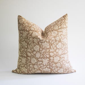 May include: A square throw pillow featuring a light tan base color with an intricate, repeating white floral and botanical pattern embroidered across the entire surface. The design includes detailed depictions of various flowers and leaves.