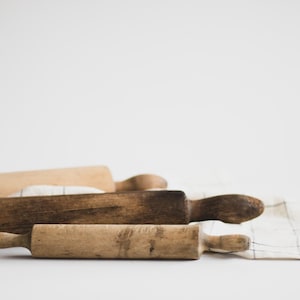 May include: Three wooden rolling pins of varying shades of brown rest on a white and beige checkered cloth. The rolling pins are of different sizes and show signs of wear, suggesting they are well-used kitchen tools.