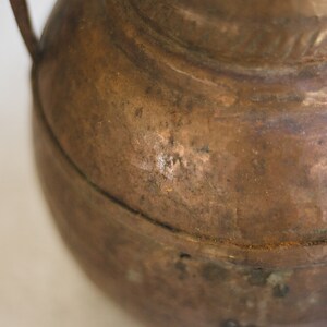 May include: A close-up of a copper pitcher with a handle. The pitcher has a worn, aged look with a patina finish.
