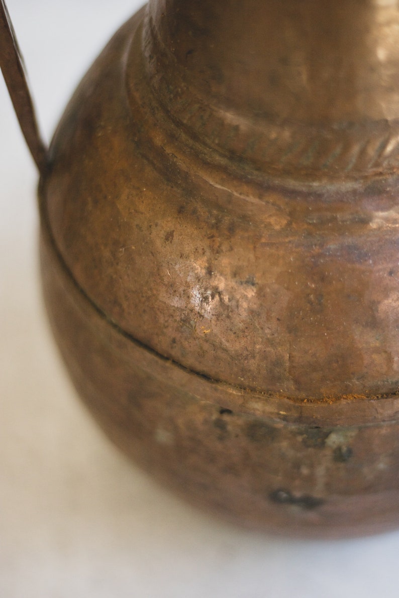 May include: A close-up of a copper pitcher with a handle. The pitcher has a worn, aged look with a patina finish.