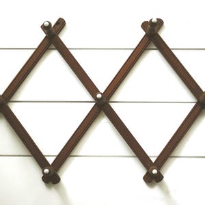 May include: An expandable wooden wall rack featuring a brown finish and a diamond-shaped lattice structure. The rack includes multiple pegs with white rounded tips, designed for hanging items, and is constructed with intersecting wooden slats.
