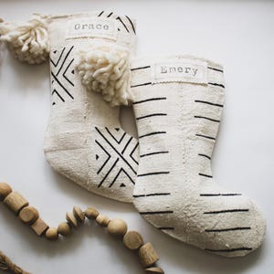 May include: Two off-white fabric Christmas stockings featuring black printed patterns. One stocking displays geometric diamond shapes and a tag reading Grace, while the other features horizontal lines and a tag reading Emery. Both have large cream yarn pom-poms.