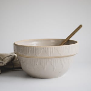 Può includere: Una ciotola in ceramica color crema con un motivo decorativo in rilievo sul bordo. Un cucchiaio di legno è all'interno della ciotola. Un panno piegato con un motivo marrone e nero è a sinistra della ciotola.