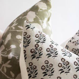 Puede incluir: Dos almohadas decorativas con patrones florales. La almohada de la izquierda tiene un fondo verde con siluetas de flores blancas. La almohada de la derecha tiene un fondo beige con un patr&oacute;n floral negro y marr&oacute;n.