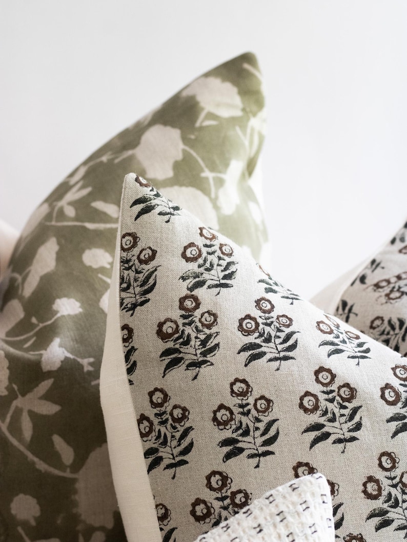 Puede incluir: Dos almohadas decorativas con patrones florales. La almohada de la izquierda tiene un fondo verde con siluetas de flores blancas. La almohada de la derecha tiene un fondo beige con un patr&oacute;n floral negro y marr&oacute;n.