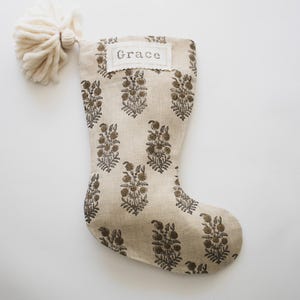 Könnte beinhalten: Ein beiger Weihnachtssocken mit braunem Blumenmuster und dem Namen "Grace" bestickt auf der Vorderseite. Der Socken hat einen weißen Bommel oben.