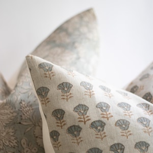 May include: A square throw cushion featuring a repeating pattern of small, light blue stylised flowers with brown stems on a beige fabric background. The cushion has a clean, piped edge and is positioned next to another cushion with a floral print.