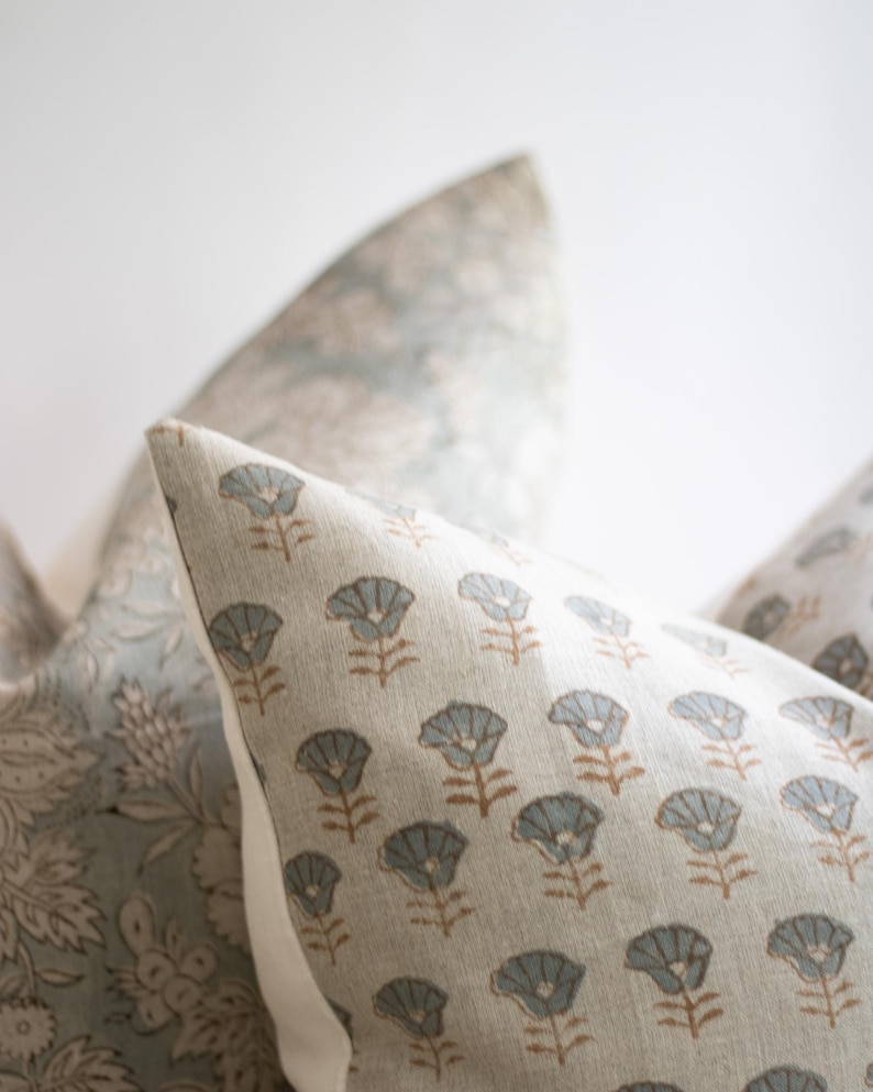 May include: A square throw cushion featuring a repeating pattern of small, light blue stylised flowers with brown stems on a beige fabric background. The cushion has a clean, piped edge and is positioned next to another cushion with a floral print.