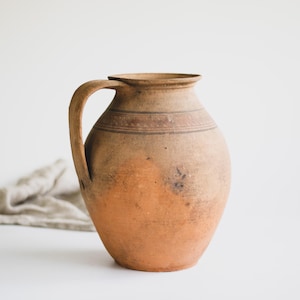 May include: A rustic, earthenware pitcher with a large handle. The pitcher has a rounded body and a narrow neck. It is primarily a warm, earthy orange-brown color, with darker brown bands around the neck. The pitcher has a vintage, antique aesthetic.