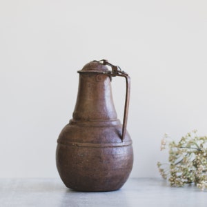 May include: A brown metal pitcher with a handle and a lid. The pitcher has a rounded body and a slightly flared top.