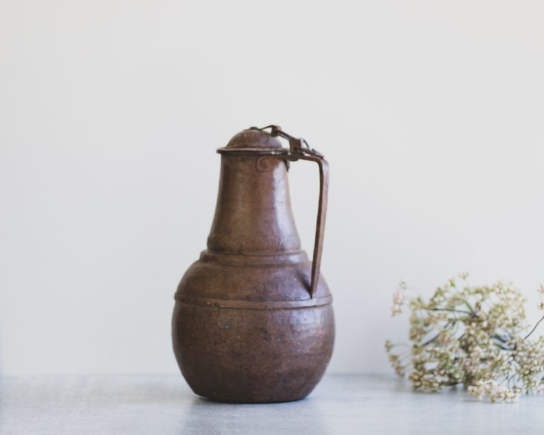 May include: A brown metal pitcher with a handle and a lid. The pitcher has a rounded body and a slightly flared top.