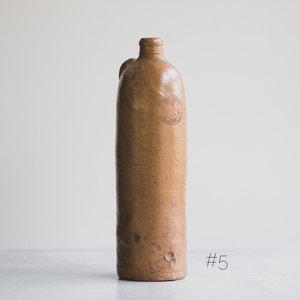 May include: An antique brown stoneware bottle with a narrow neck and a small handle. The bottle has a slightly textured surface and a circular embossed design near the top. This vintage bottle was likely used for storing liquids.